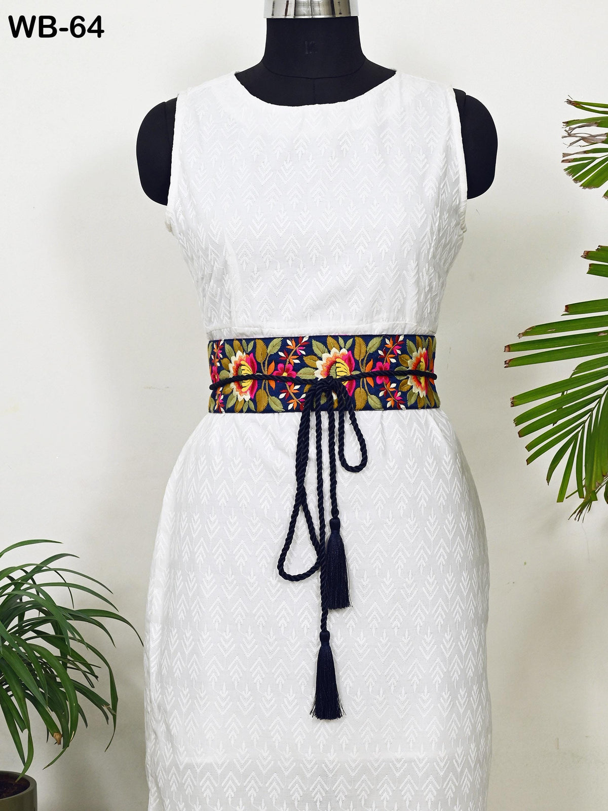 A mannequin wearing a white sleeveless dress with a colorful floral belt, placed against a white wall with a potted plant in the background.