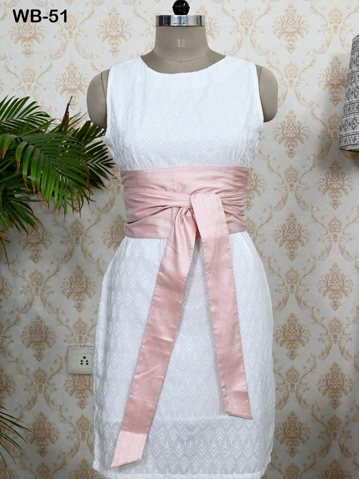 A white dress with a pink sash, displayed on a mannequin against a patterned wallpaper.