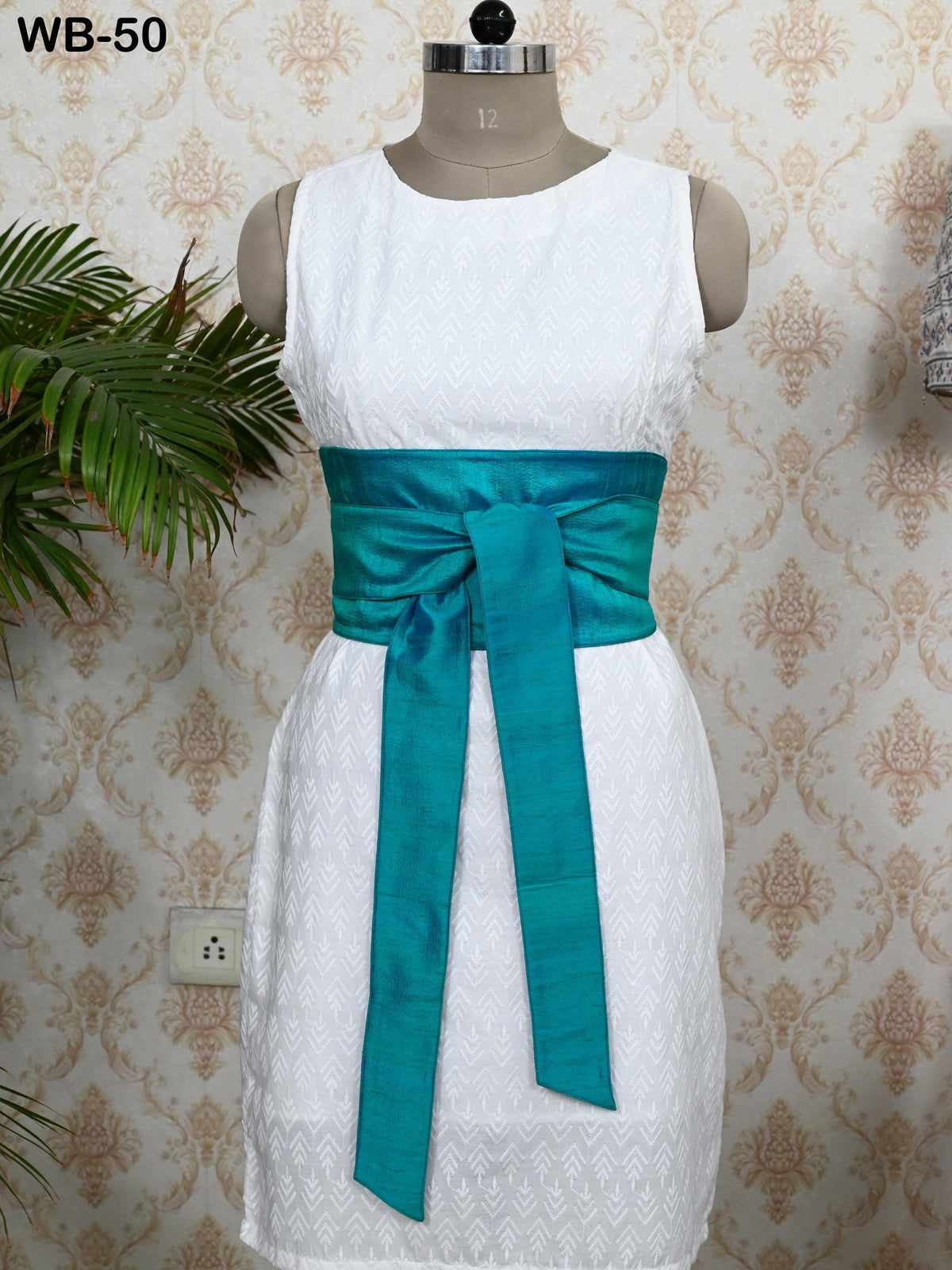 A mannequin wearing a white dress with a turquoise sash, displayed against a patterned wallpaper.