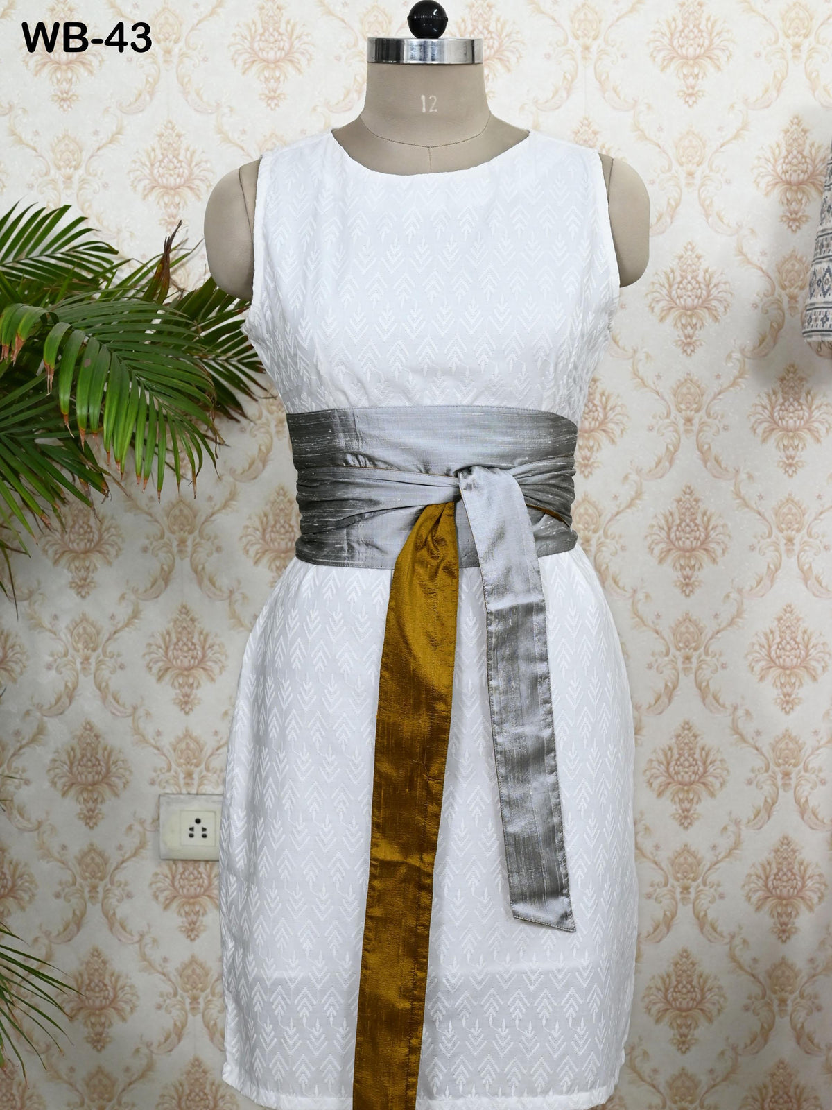 A mannequin wearing a white sleeveless dress with a grey sash tied around the waist. The dress has a gold and silver belt, and there is a plant visible in the background.