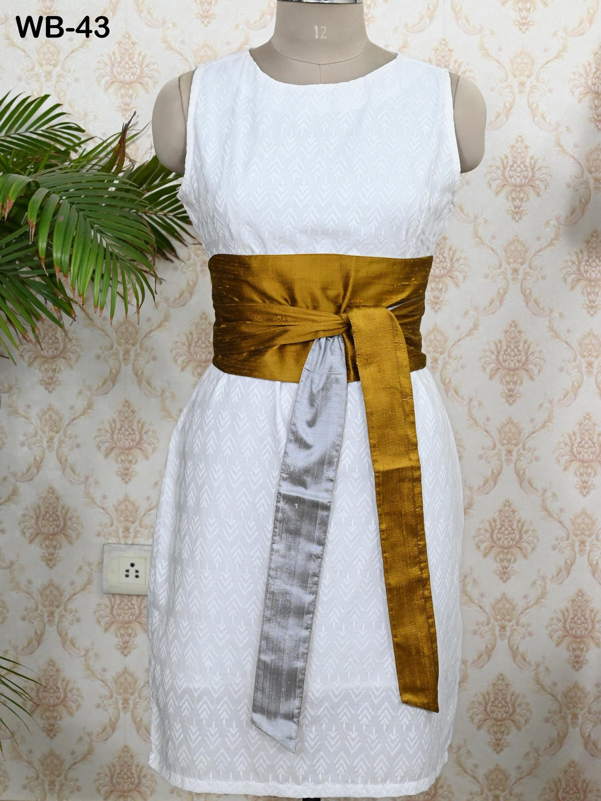 A white dress with a gold sash, displayed on a mannequin against a patterned wallpaper.