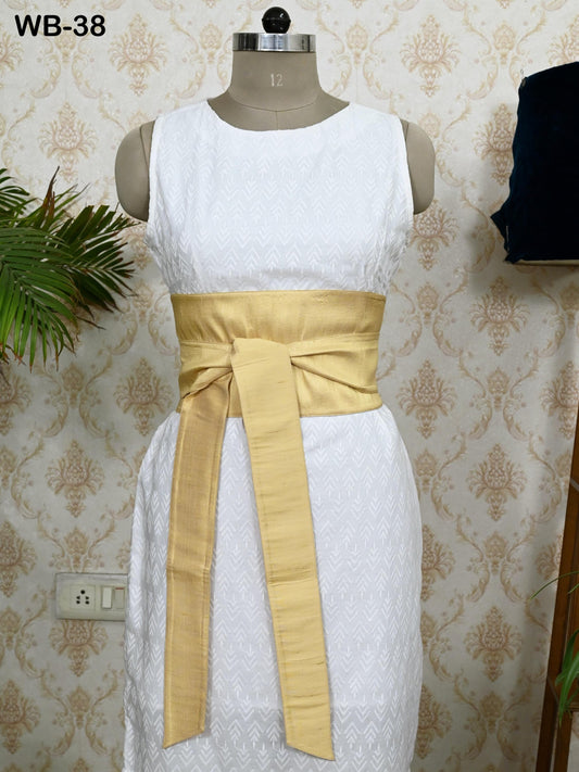 A white dress with a gold sash, displayed on a mannequin against a patterned wallpaper.