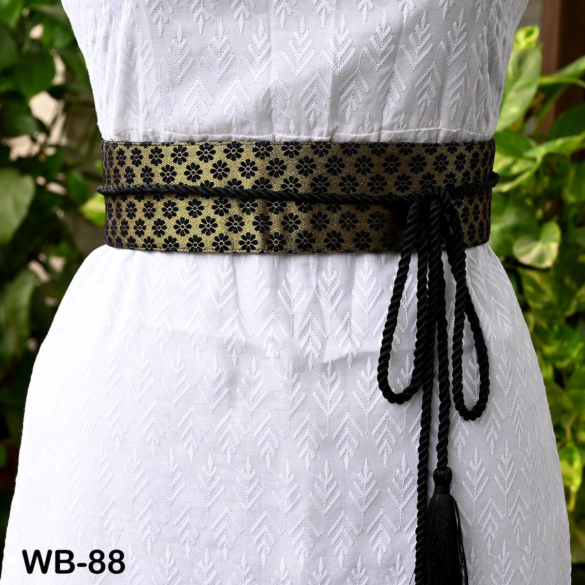 A white dress with a black and gold patterned belt, featuring a tassel at the end.