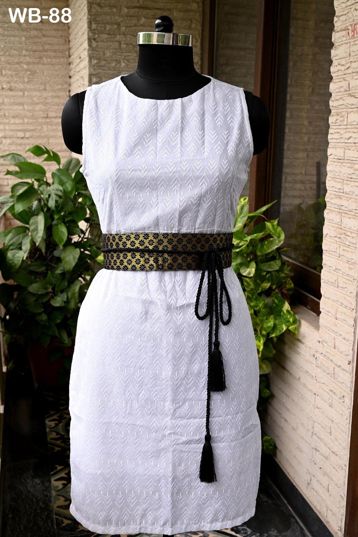 A white sleeveless dress with a black and gold belt, displayed on a mannequin in front of a brick wall with plants.