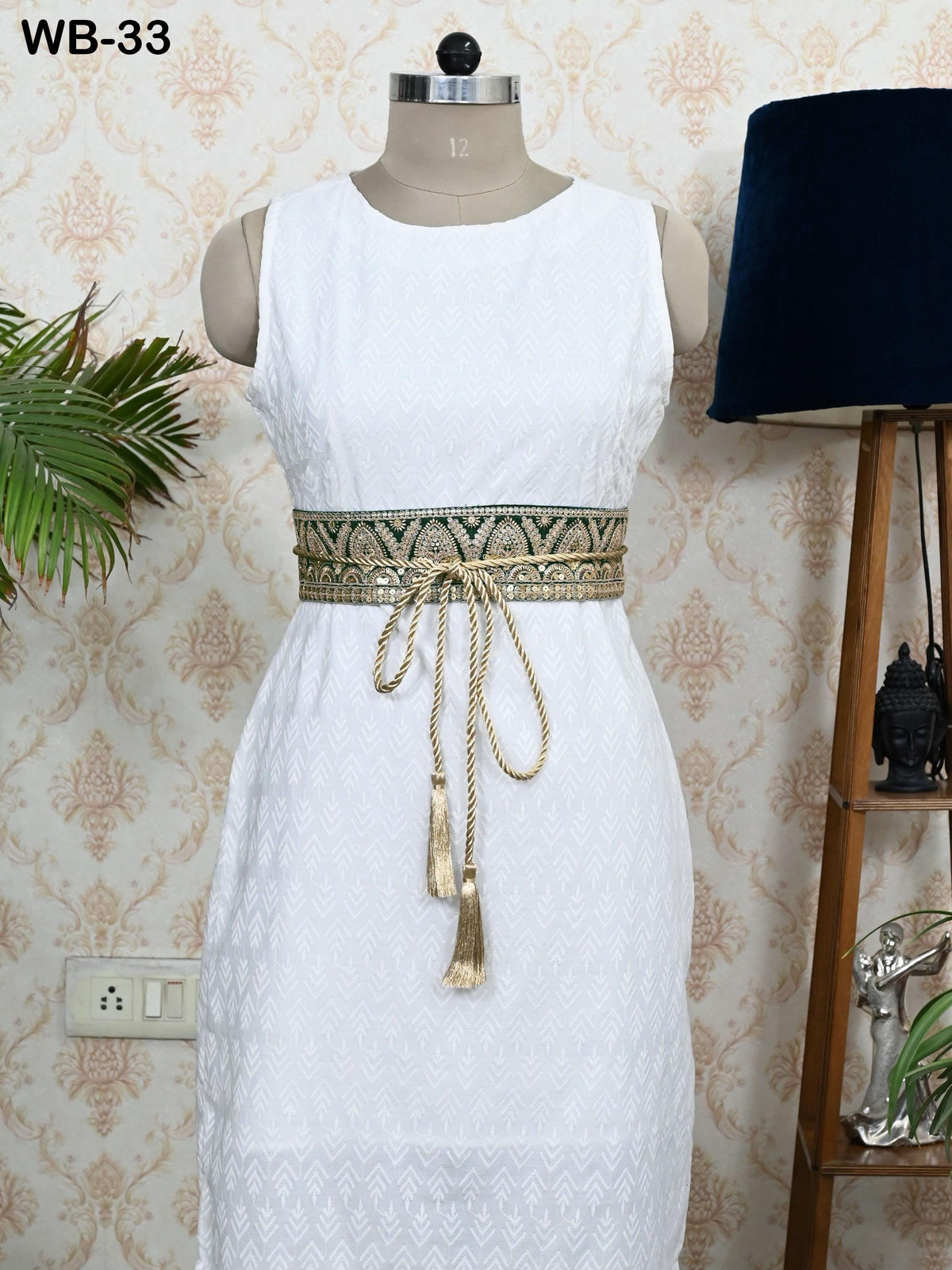 A white dress with a gold belt and tassels is displayed on a mannequin, placed on a wooden stand next to a plant and a lamp.