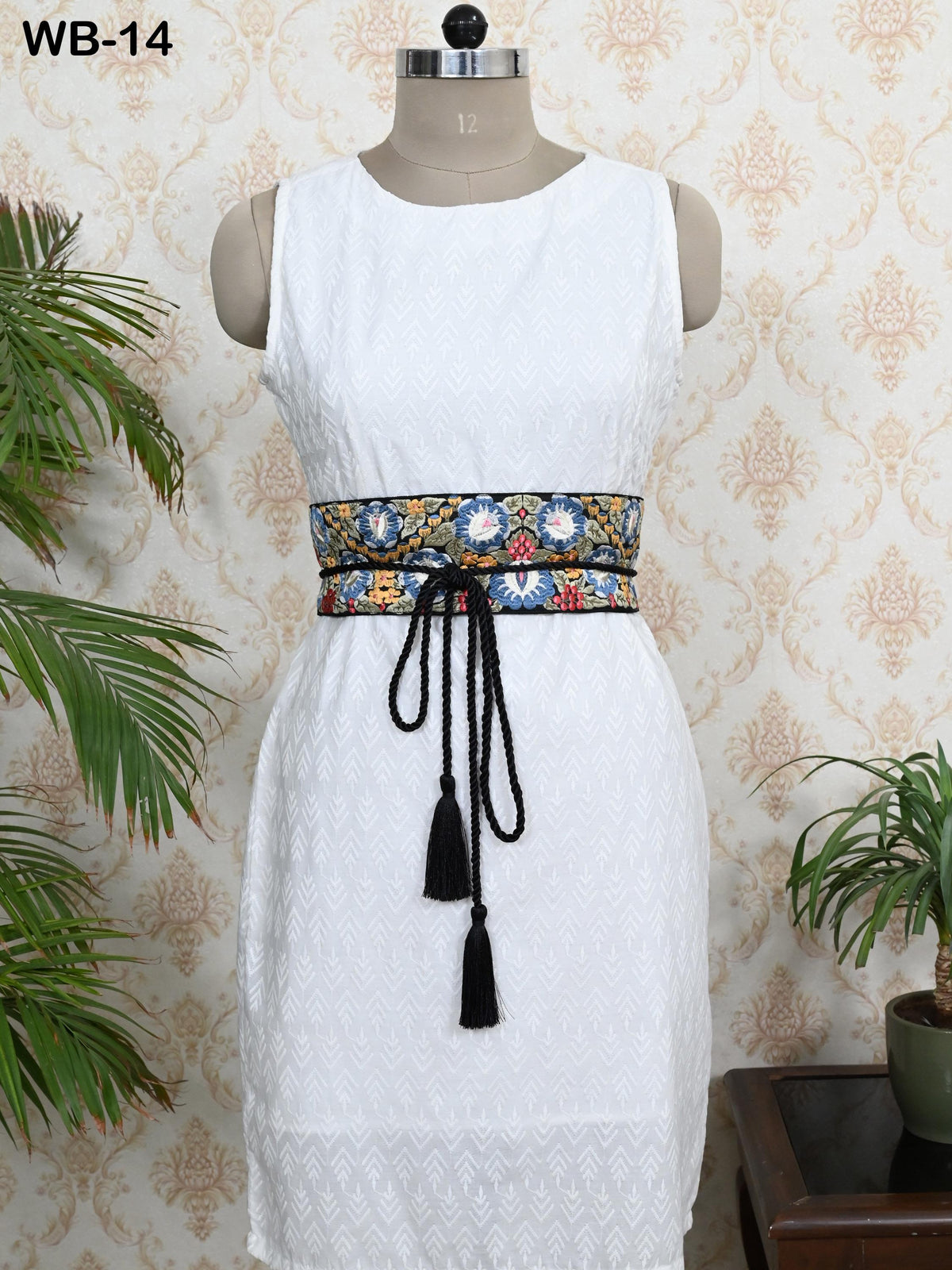 A mannequin wearing a white sleeveless dress with a decorative belt featuring a floral pattern and tassels. The dress is displayed against a patterned wallpaper background, with a potted plant visible to the right.