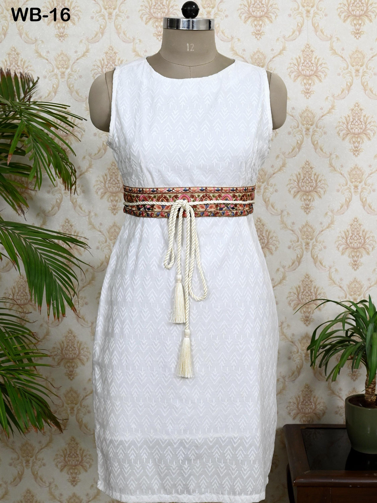 A white sleeveless dress with a decorative belt, displayed on a mannequin against a patterned wallpaper background.