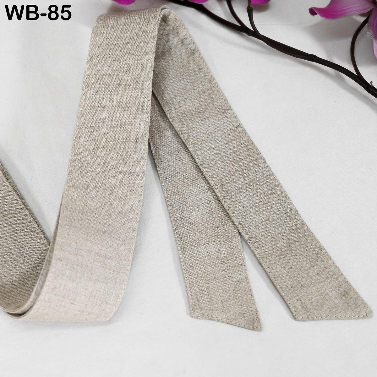 A beige fabric ribbon is lying on a white surface, with a branch of purple flowers visible in the background.