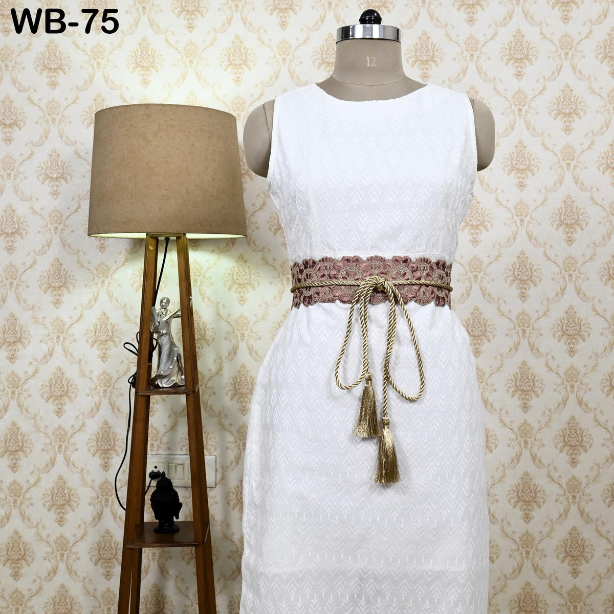 A white dress with a decorative belt, displayed on a mannequin, next to a lamp and a small statue on a wooden stand.