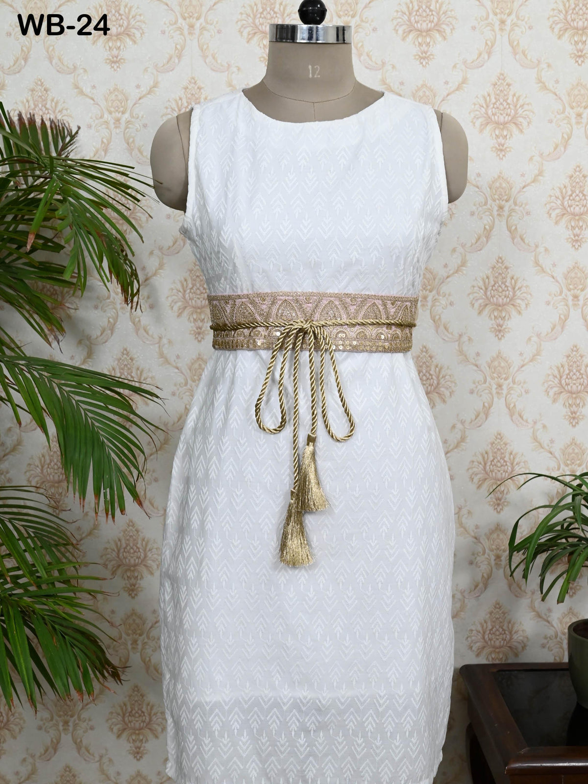 A white sleeveless dress with a gold belt and tassels, displayed on a mannequin against a patterned wallpaper.