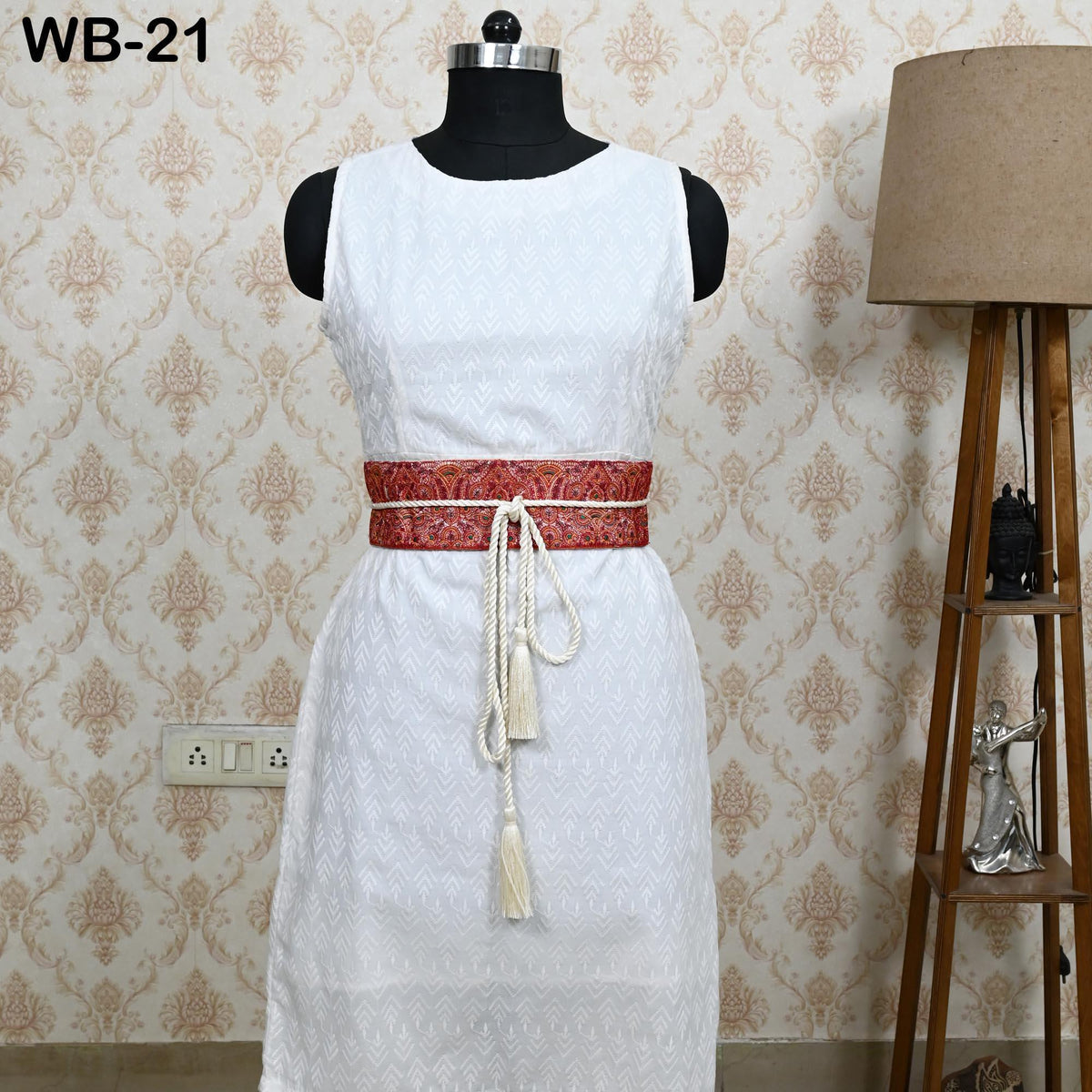A white dress with a red belt and tassels is displayed on a mannequin, placed against a wall with a lamp and a patterned wallpaper.