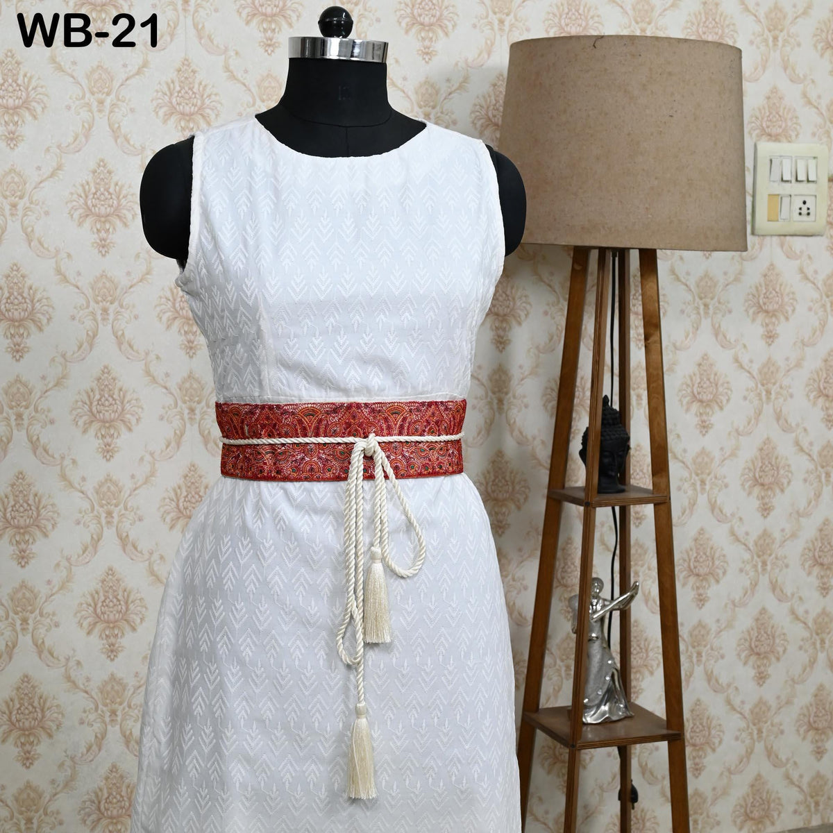 A mannequin wearing a white sleeveless dress with a red and white patterned belt, placed next to a wooden floor lamp.