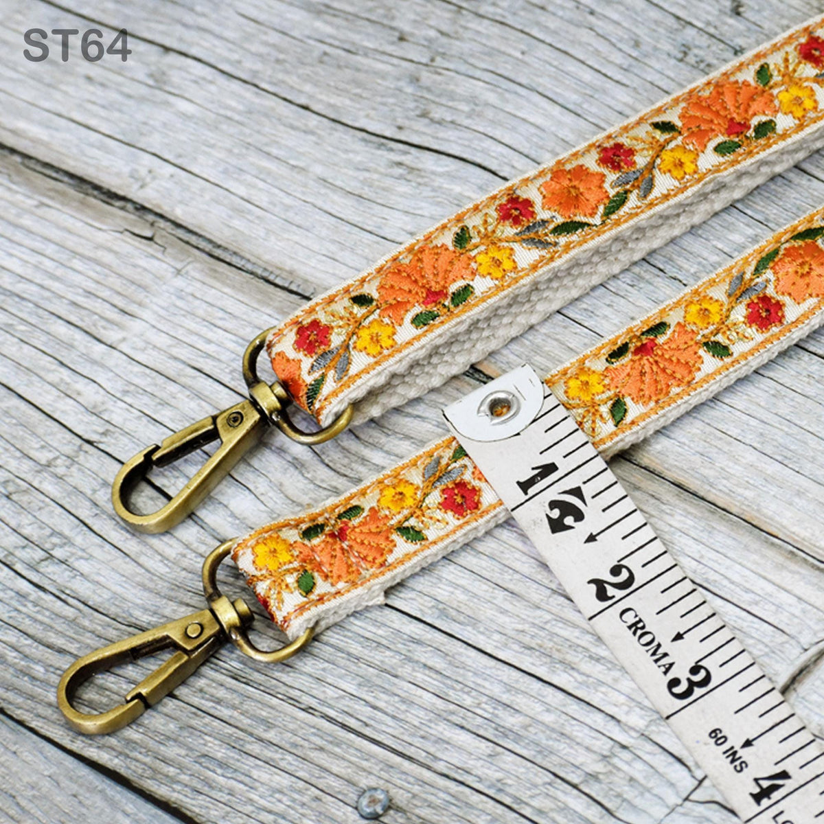 a pair of gold-colored metal straps with a floral pattern, resting on a wooden surface. A ruler is placed next to the straps, indicating the size or length of the straps.