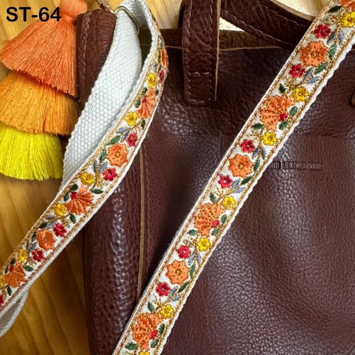 A brown leather handbag with a decorative floral embroidered strap and a white tassel.
