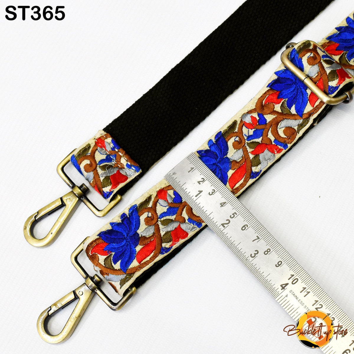 a black strap with a floral pattern and two metal hooks, one of which is attached to a ruler for size comparison.