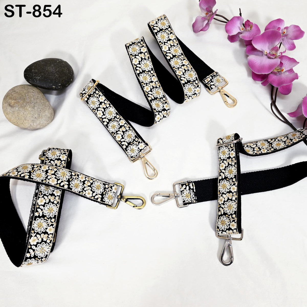a collection of black and white floral straps arranged on a white background. The straps are adorned with gold hardware and are accompanied by a few rocks and purple orchids.