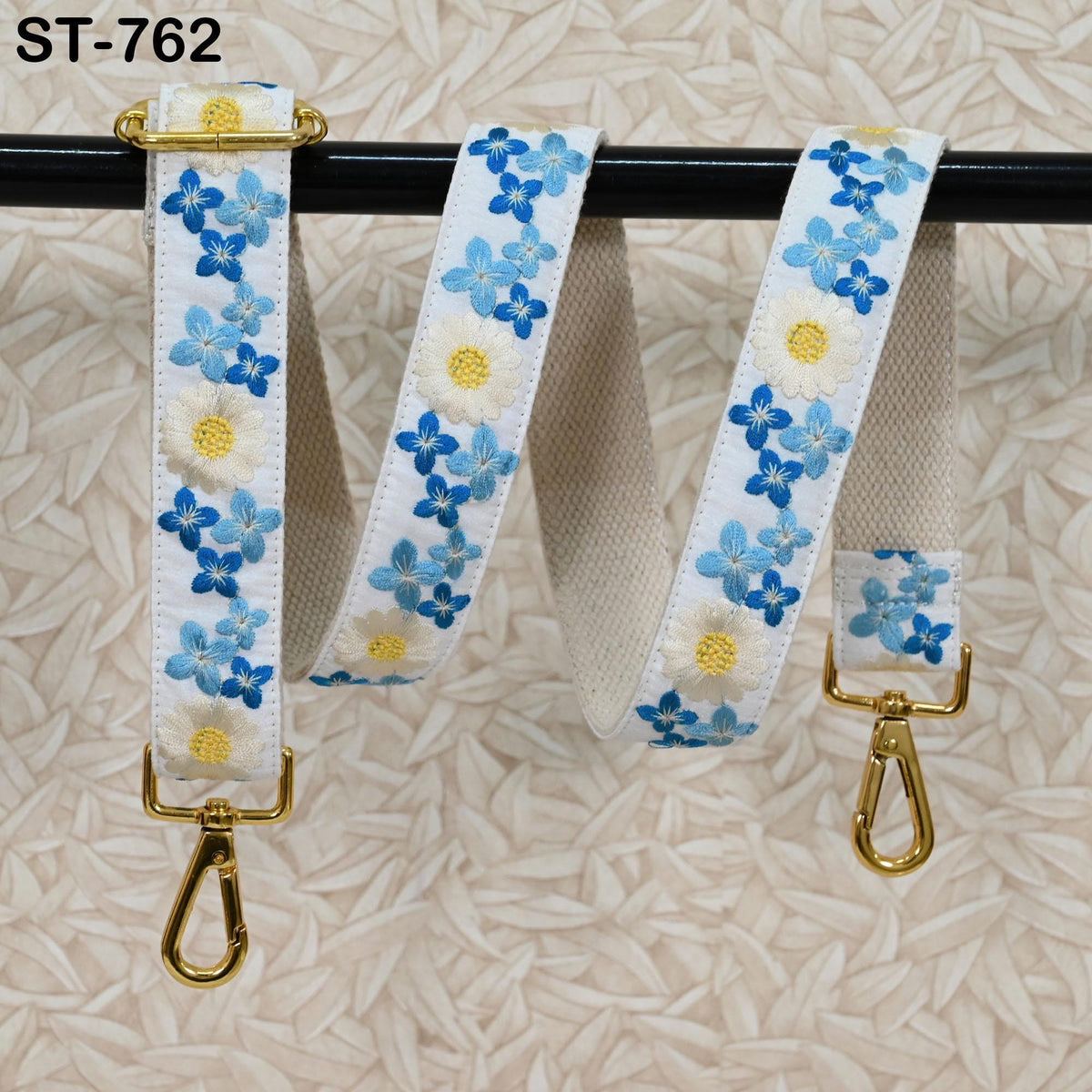 A white strap with blue and yellow floral embroidery, featuring a gold metal clasp and hook, is displayed against a textured beige background.