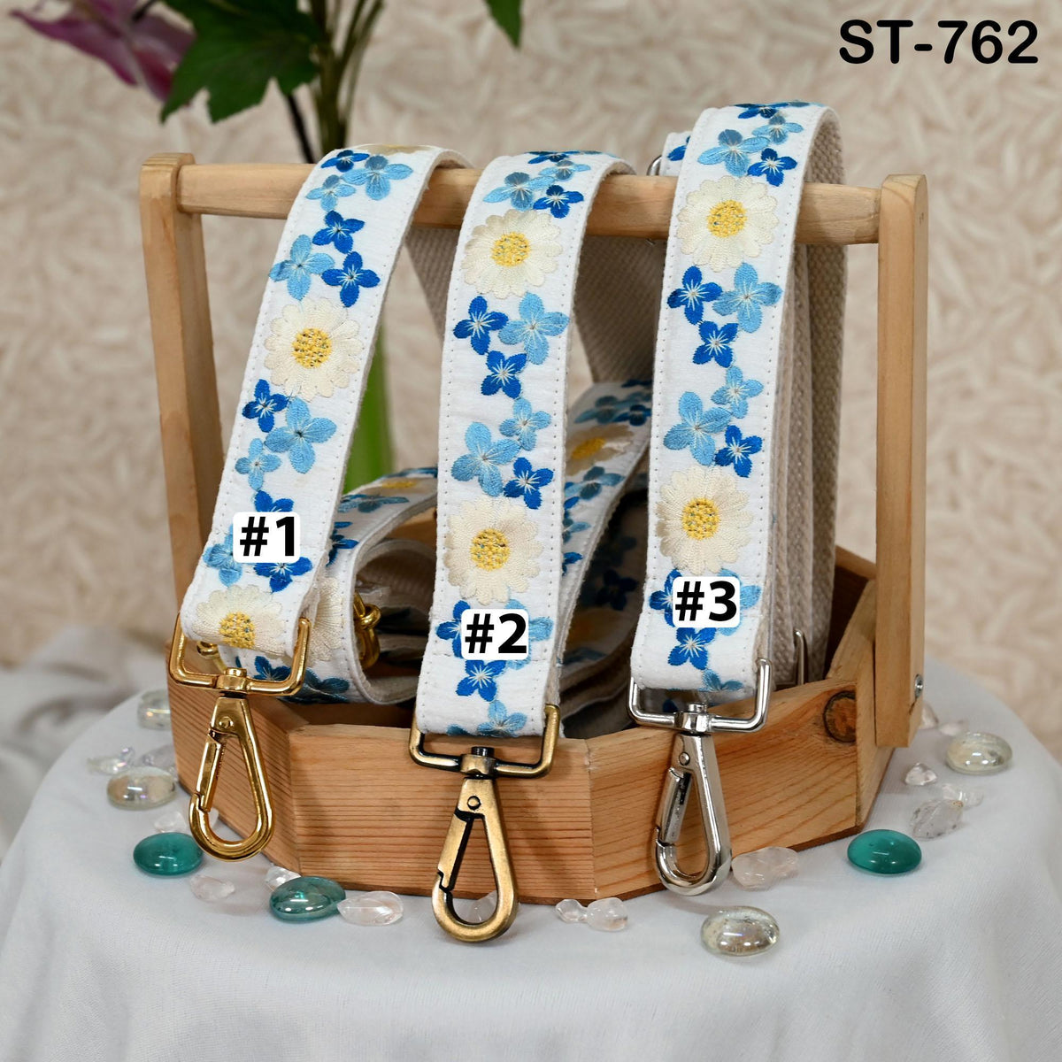 A wooden display case containing three white straps with blue flower patterns, each with a metal clasp and hook. The straps are arranged in a row, and there are small stones on the table beneath the display case.