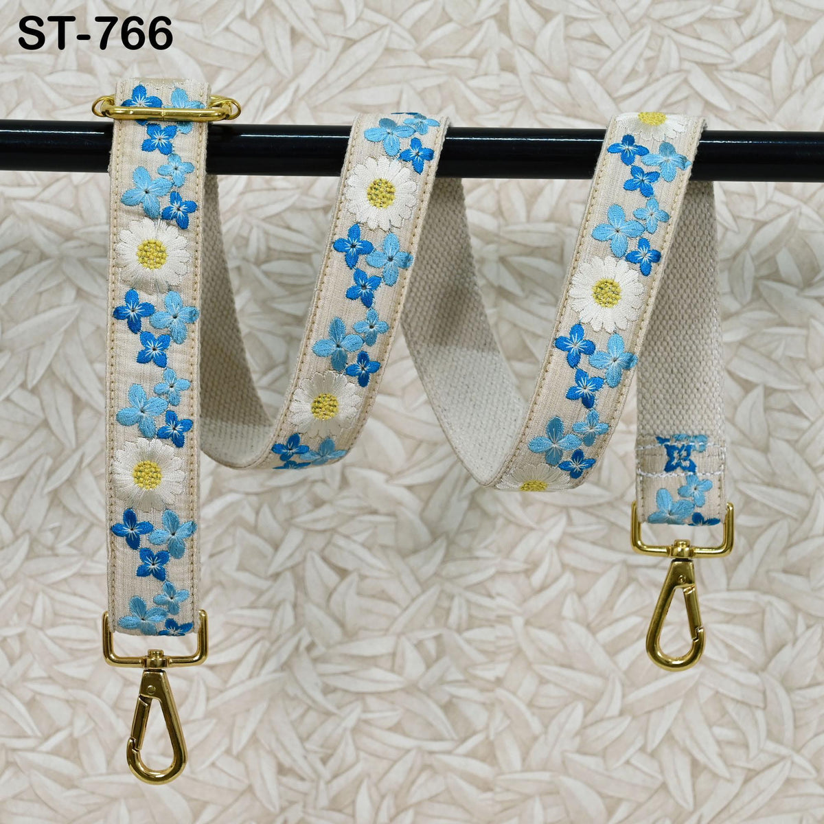 A floral-patterned strap with gold-colored metal hooks attached to it, hanging on a black rod against a white background.