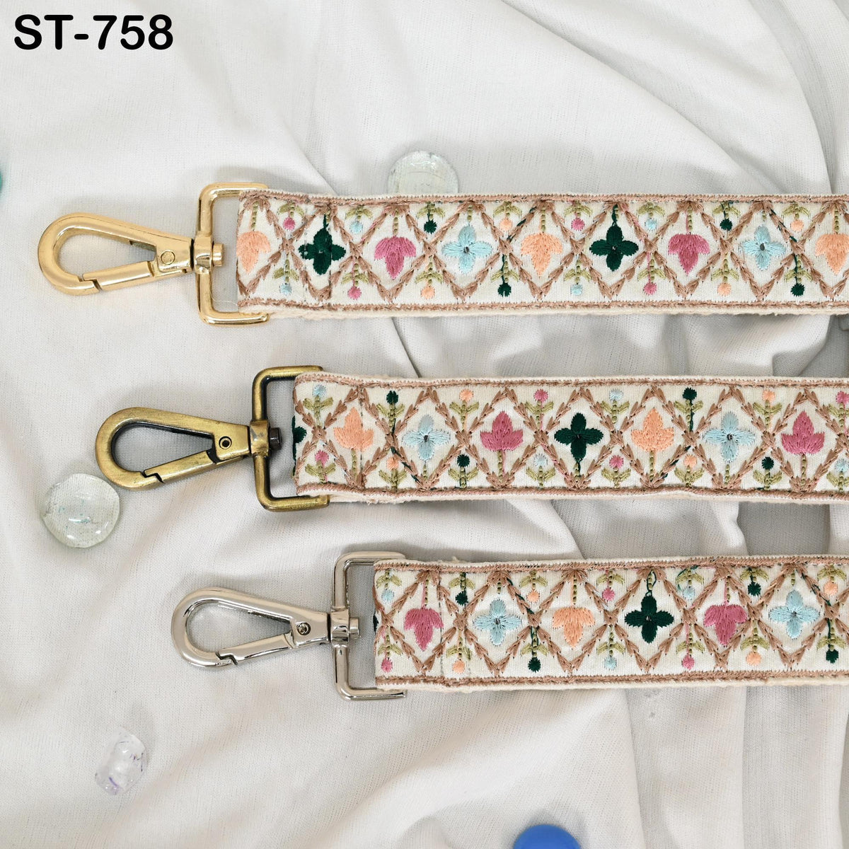 three colorful straps with intricate embroidered patterns, each with a metal clasp and hook. The straps are arranged on a white fabric background, with small decorative stones scattered around them.