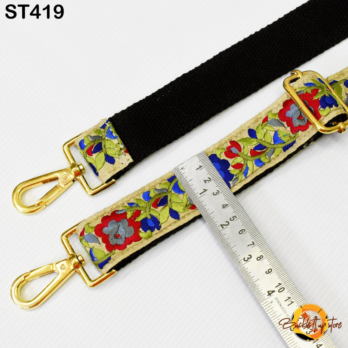 a black strap with a floral pattern and two gold-colored metal hooks, placed next to a ruler for size comparison.