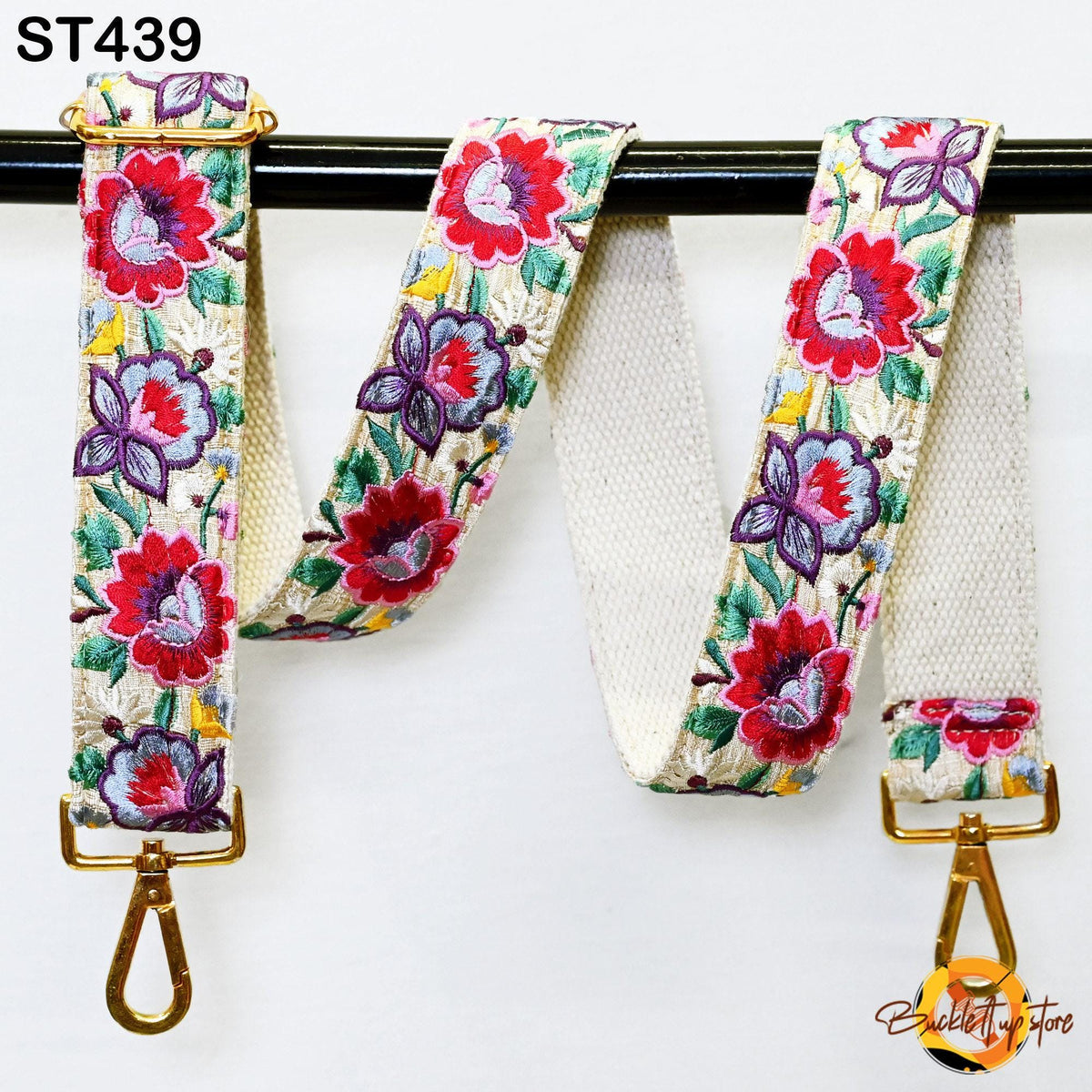 A floral embroidered strap with a gold metal buckle, hanging on a black bar.