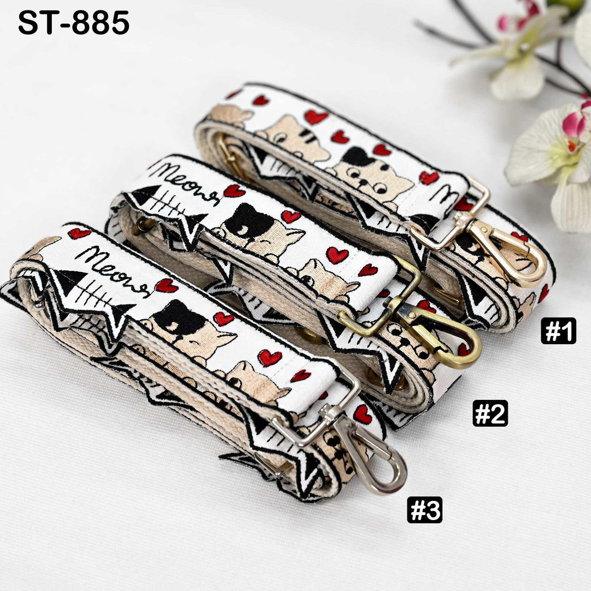 a collection of three cat-themed straps, each with a unique design and color scheme. The straps are arranged in a row, with the first one being the largest and the third one being the smallest. The straps are displayed against a white background, and there are small white flowers visible in the background.