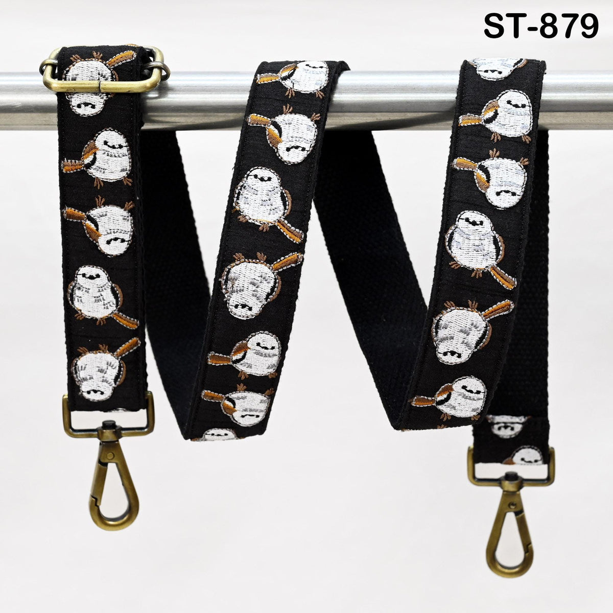 a black strap with a repeating pattern of white birds on it. The strap has a brass buckle and a gold hook, and it is hanging from a metal bar.