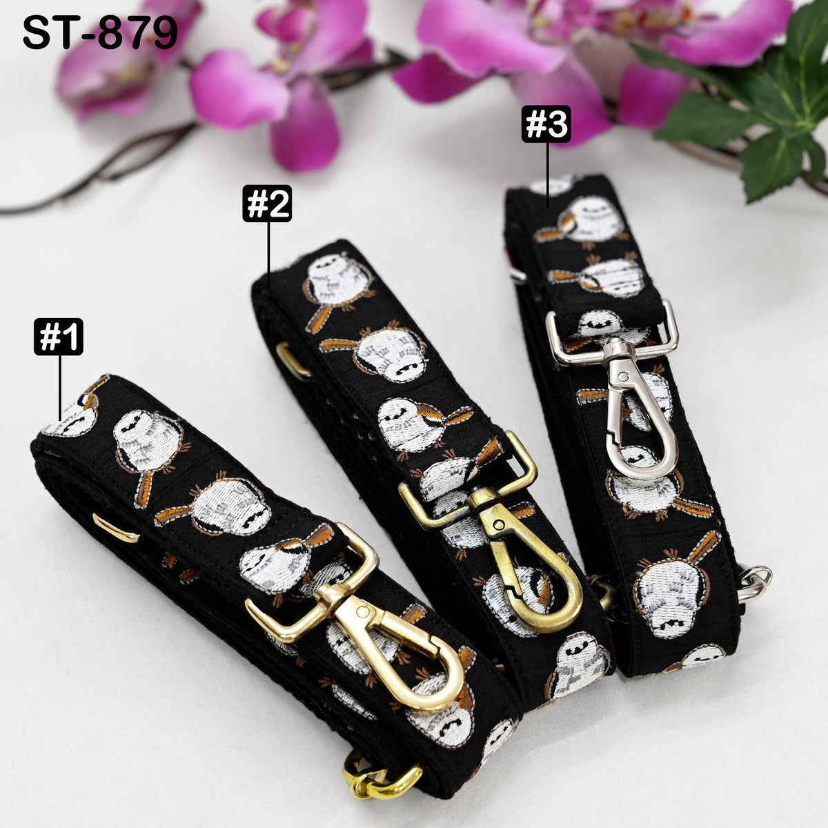 three black straps with gold buckles, each adorned with a pattern of white and black owls. The straps are arranged in a row, with the first one on the left, the second one in the middle, and the third one on the right. The background of the image is a white surface, and there are purple flowers visible in the top right corner.