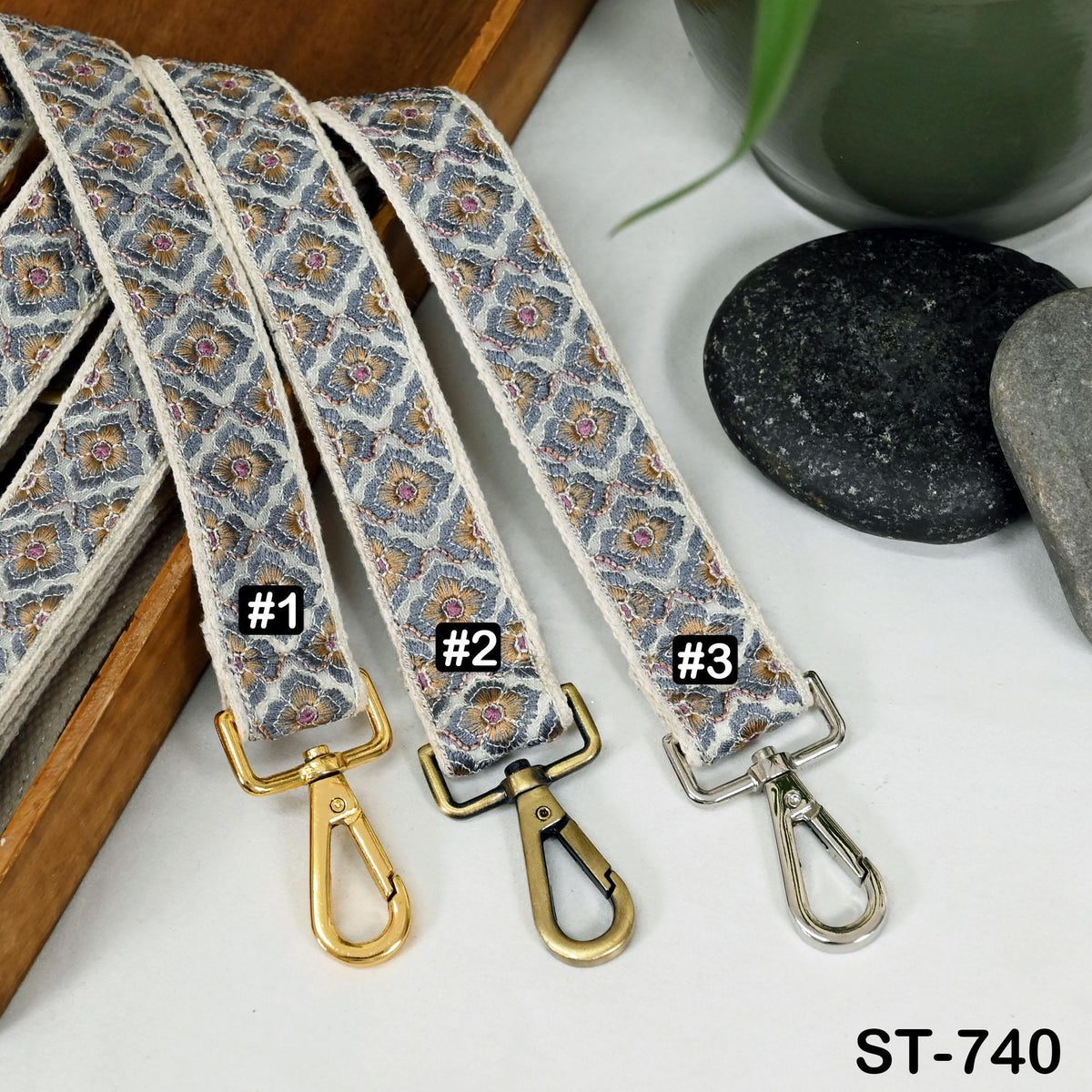 A set of three patterned straps with metal hooks, each labeled with a number, placed on a wooden surface next to a black rock and a green plant.