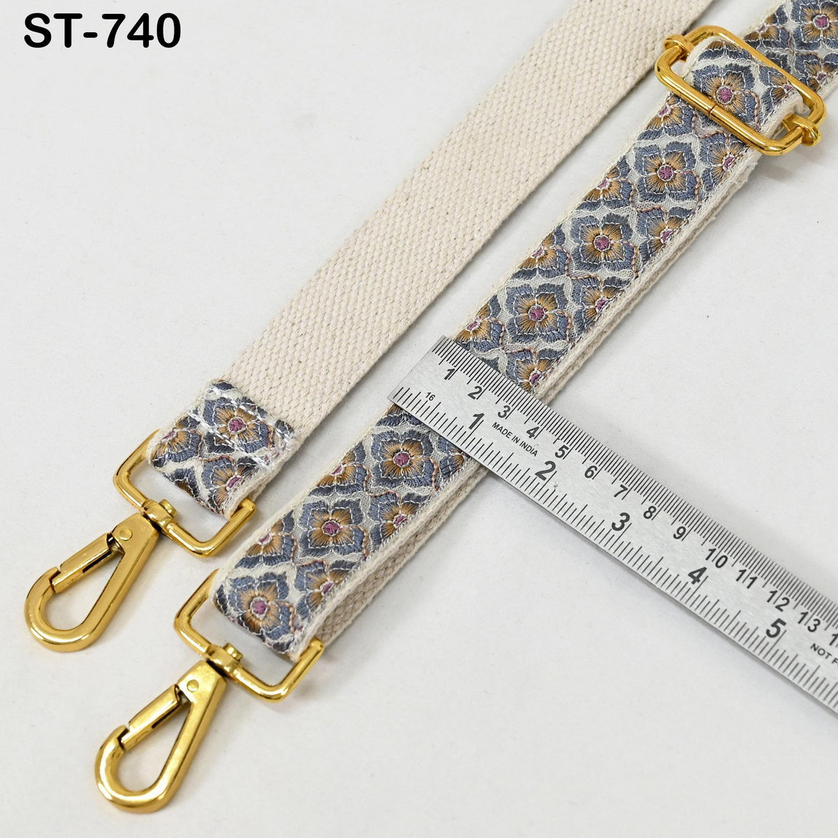 a beige strap with a floral pattern and two gold metal hooks, placed next to a ruler for size comparison.