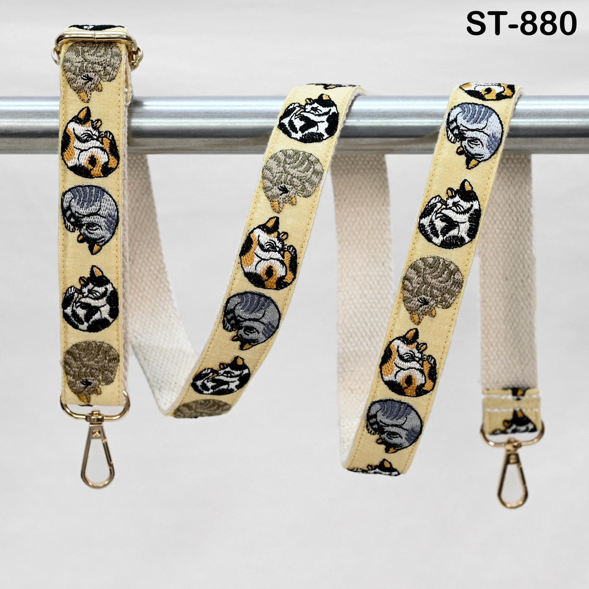a beige strap with a repeating pattern of cats, including a sleeping cat, a standing cat, and a curled-up cat, all in shades of gray and black. The strap is hanging on a metal bar, and there are two gold-colored metal hooks attached to the strap.