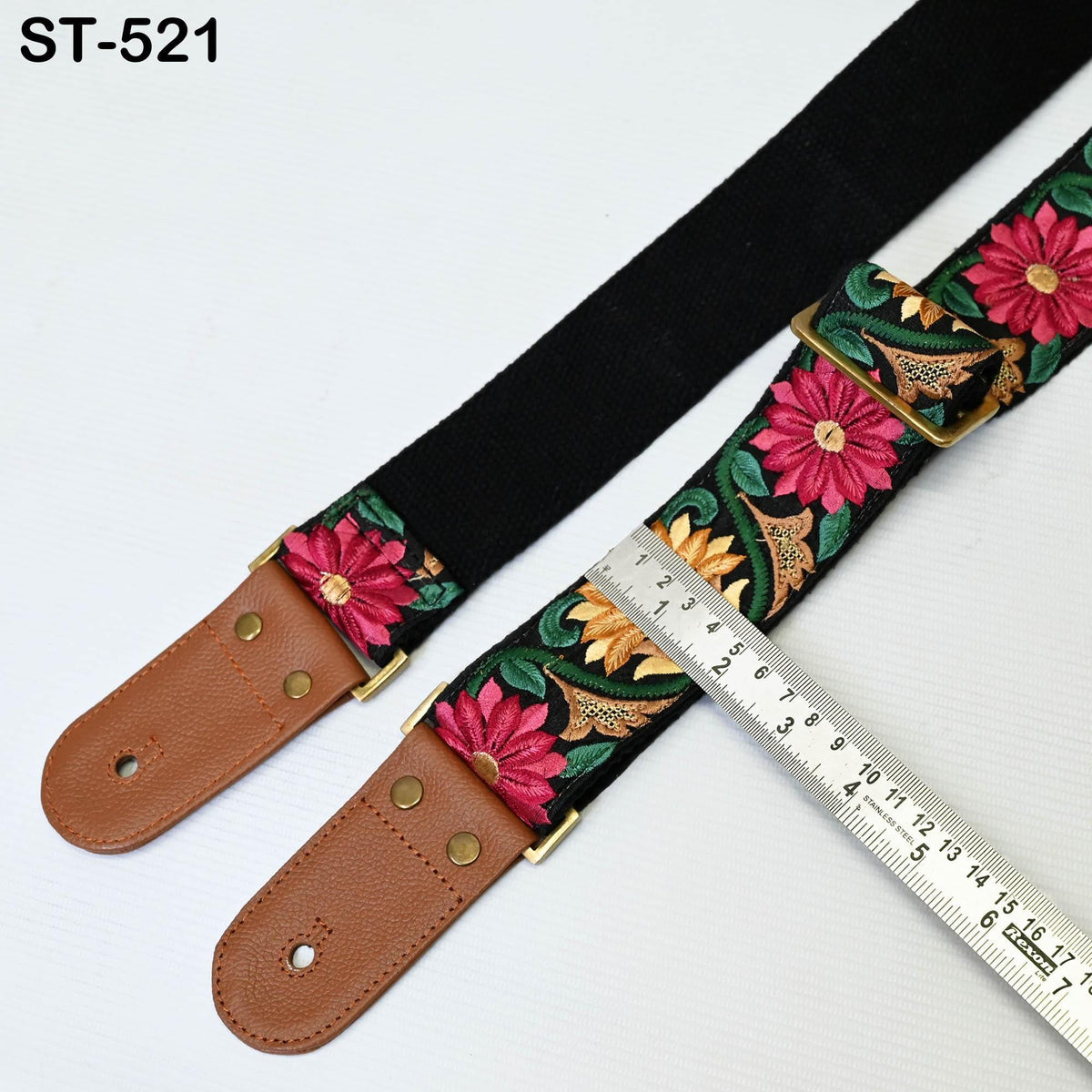 A black guitar strap with a floral pattern and a brown leather strap with a floral pattern are placed next to a metal ruler on a white background.