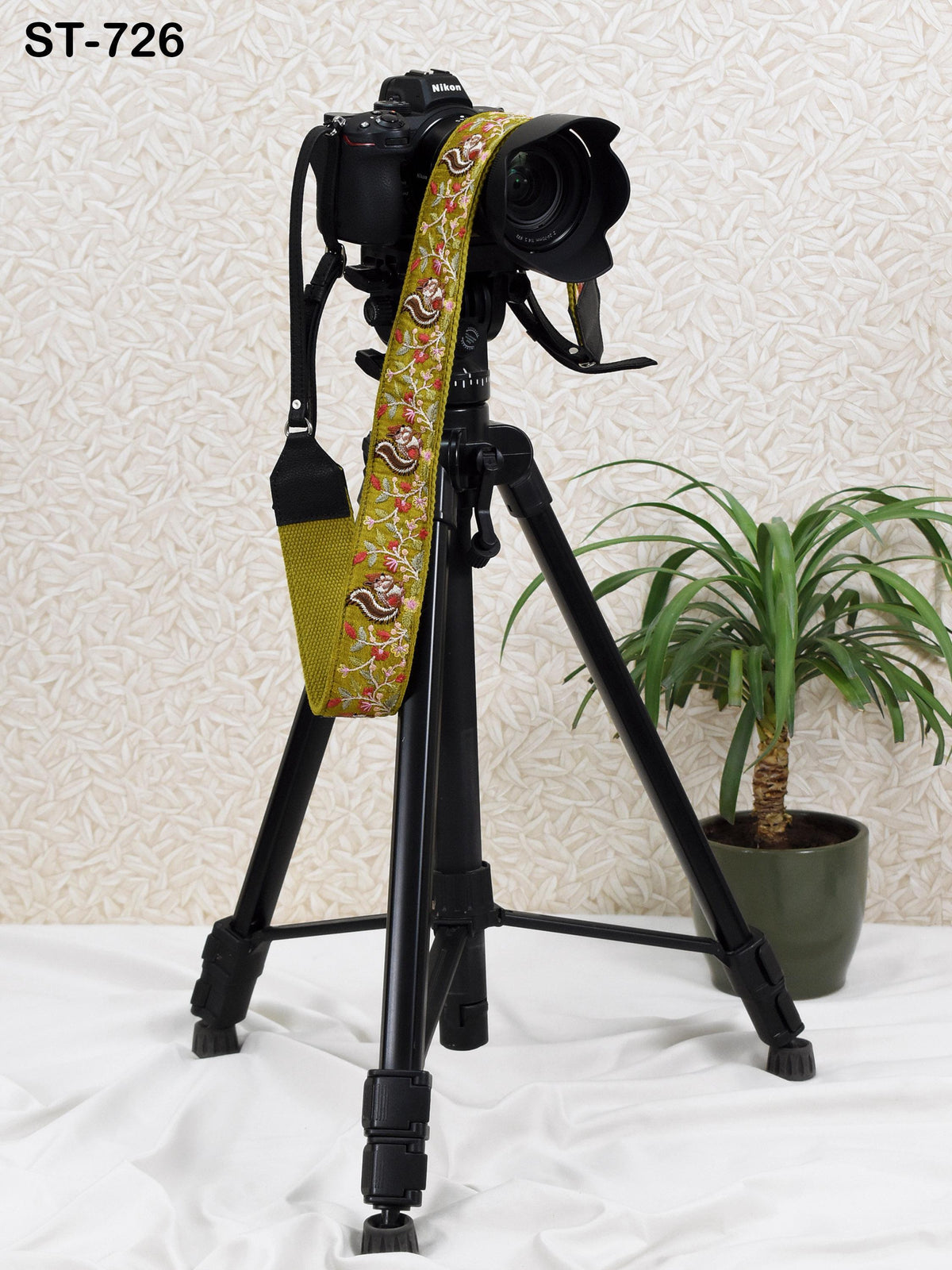 A black tripod with a camera attached to it, placed on a white surface next to a potted plant.