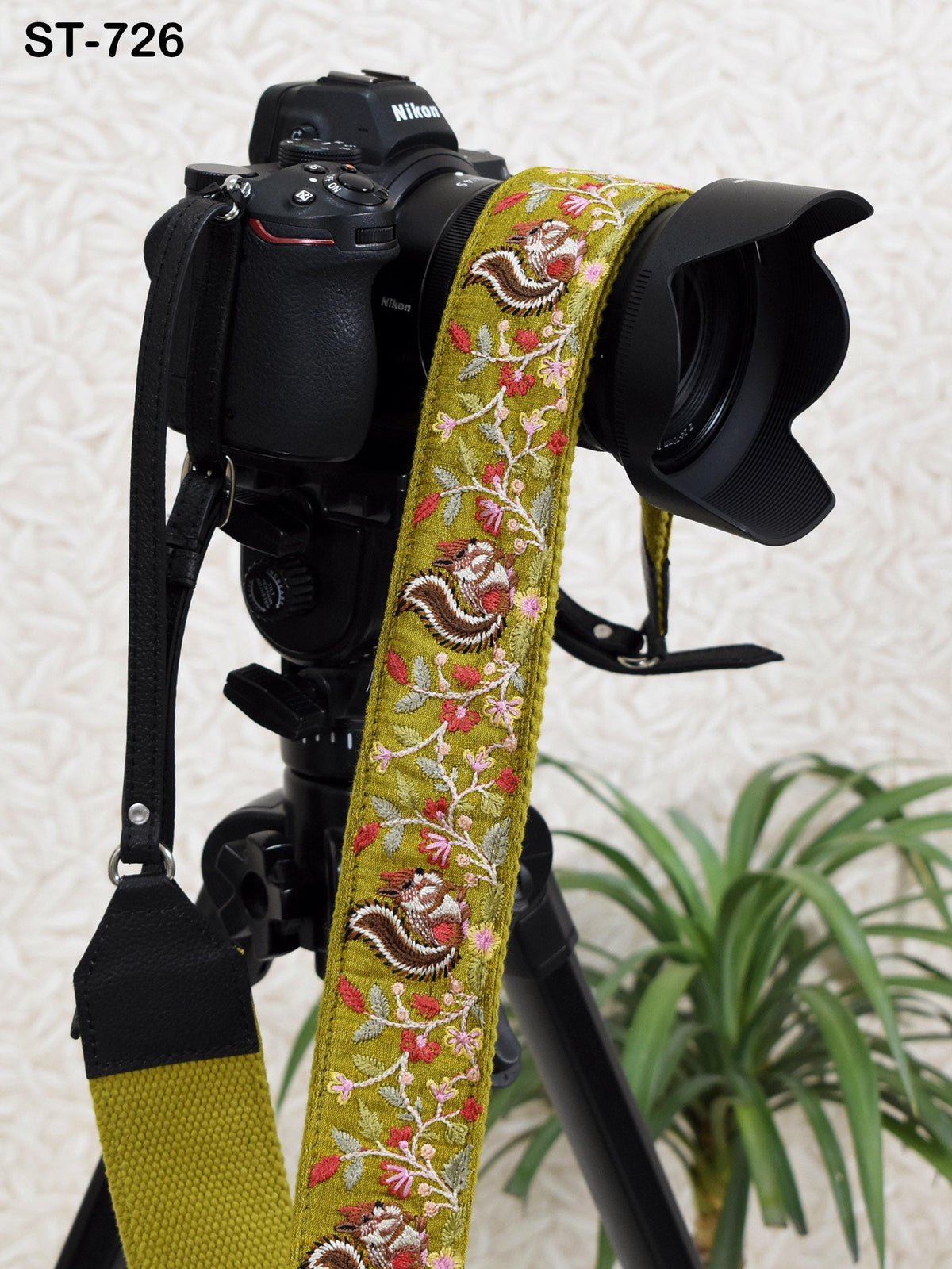 A black camera with a yellow and green floral patterned strap attached to it, placed on a tripod in front of a plant.