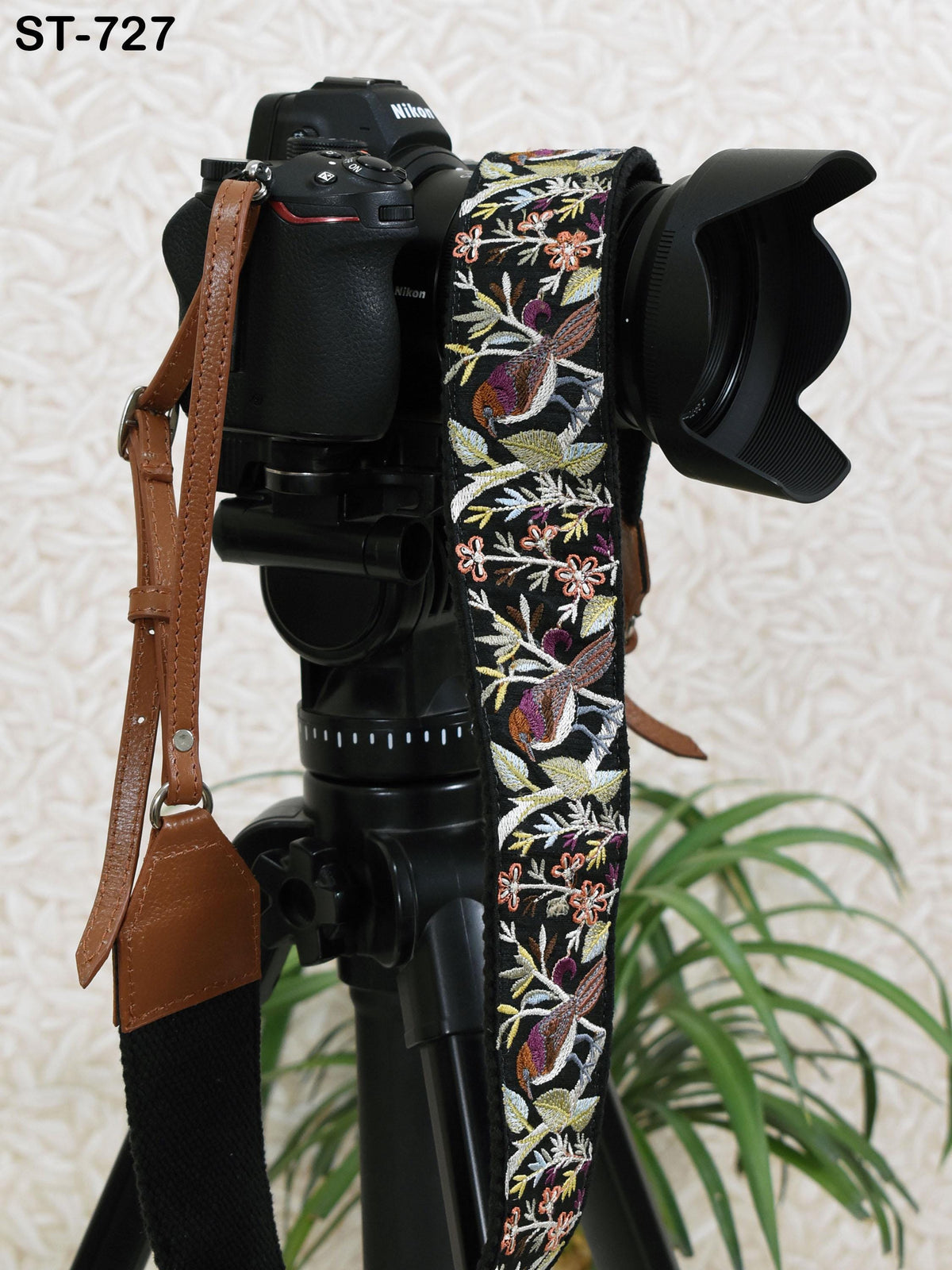 A black camera with a floral patterned strap is placed on a tripod, with a potted plant visible in the background.