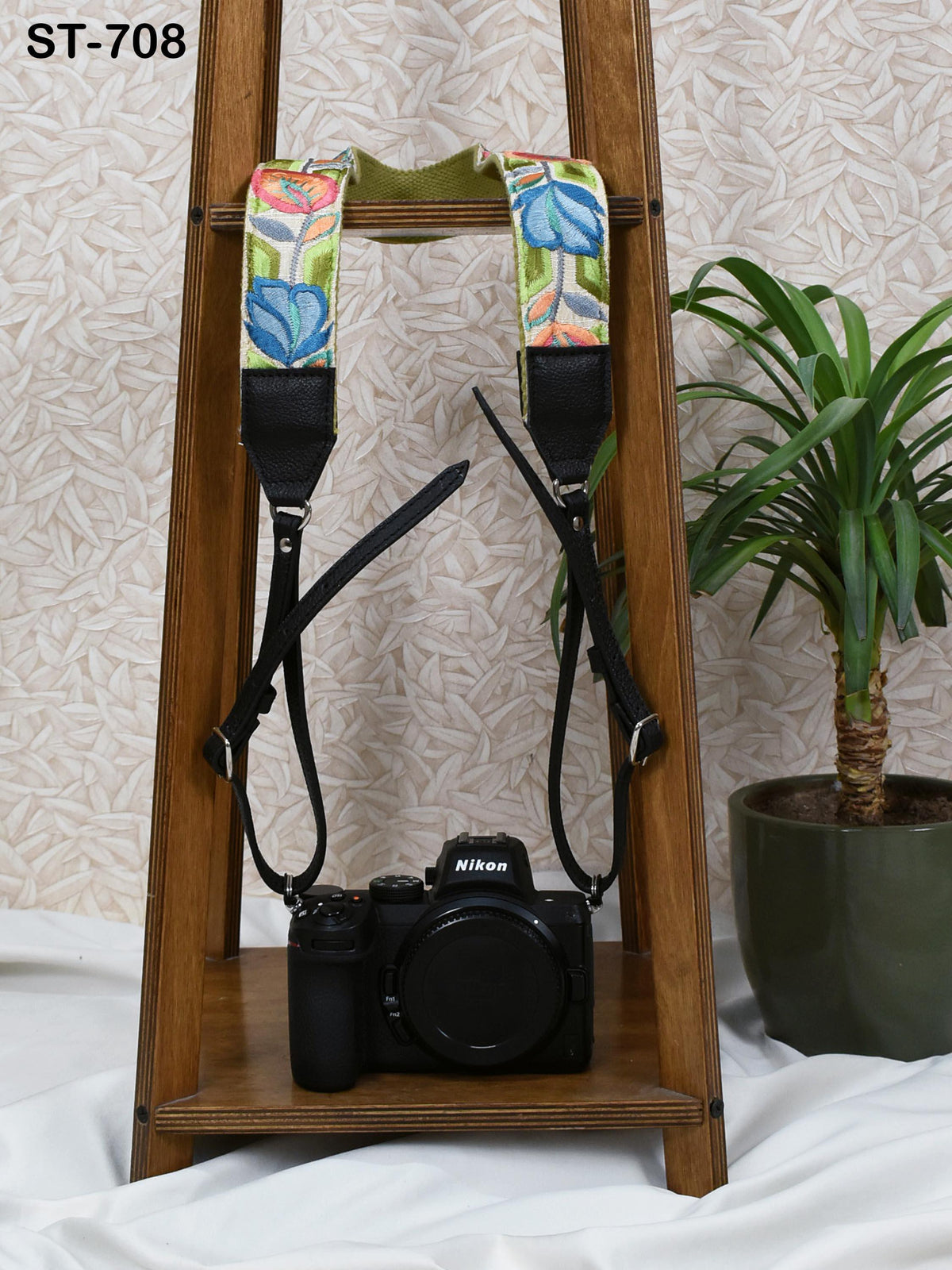 A black camera with a colorful strap is placed on a wooden stand, with a potted plant visible in the background.