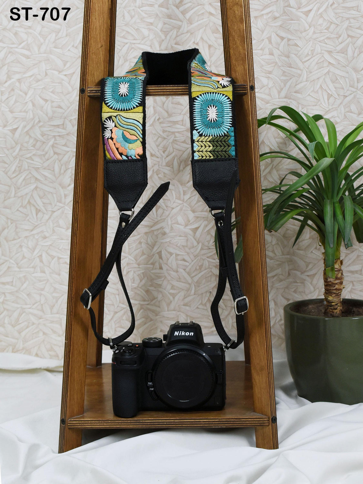 A black camera with a colorful strap is displayed on a wooden stand, with a potted plant in the background.