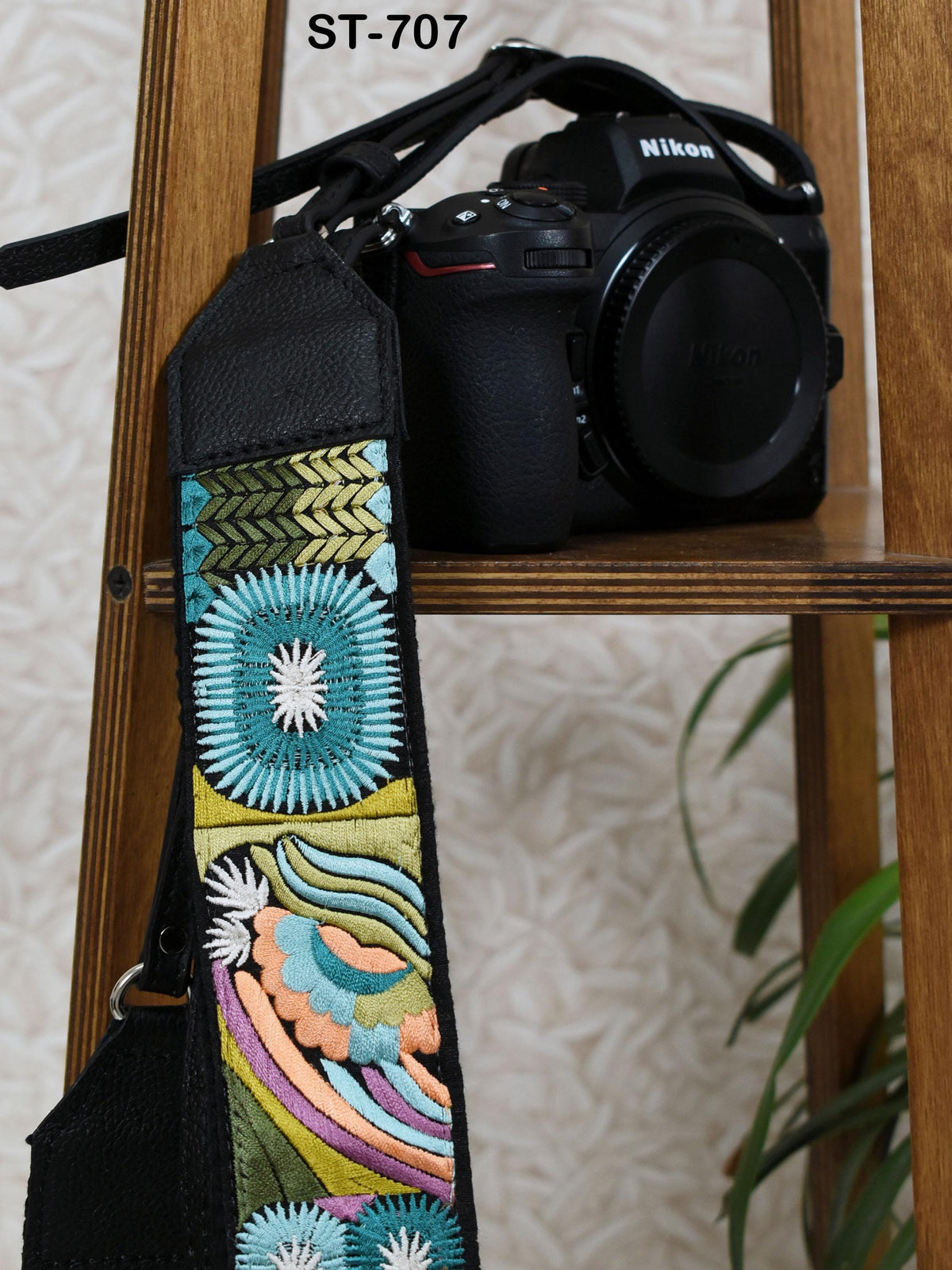 A black camera strap with colorful floral embroidery is hanging on a wooden shelf next to a Nikon camera.