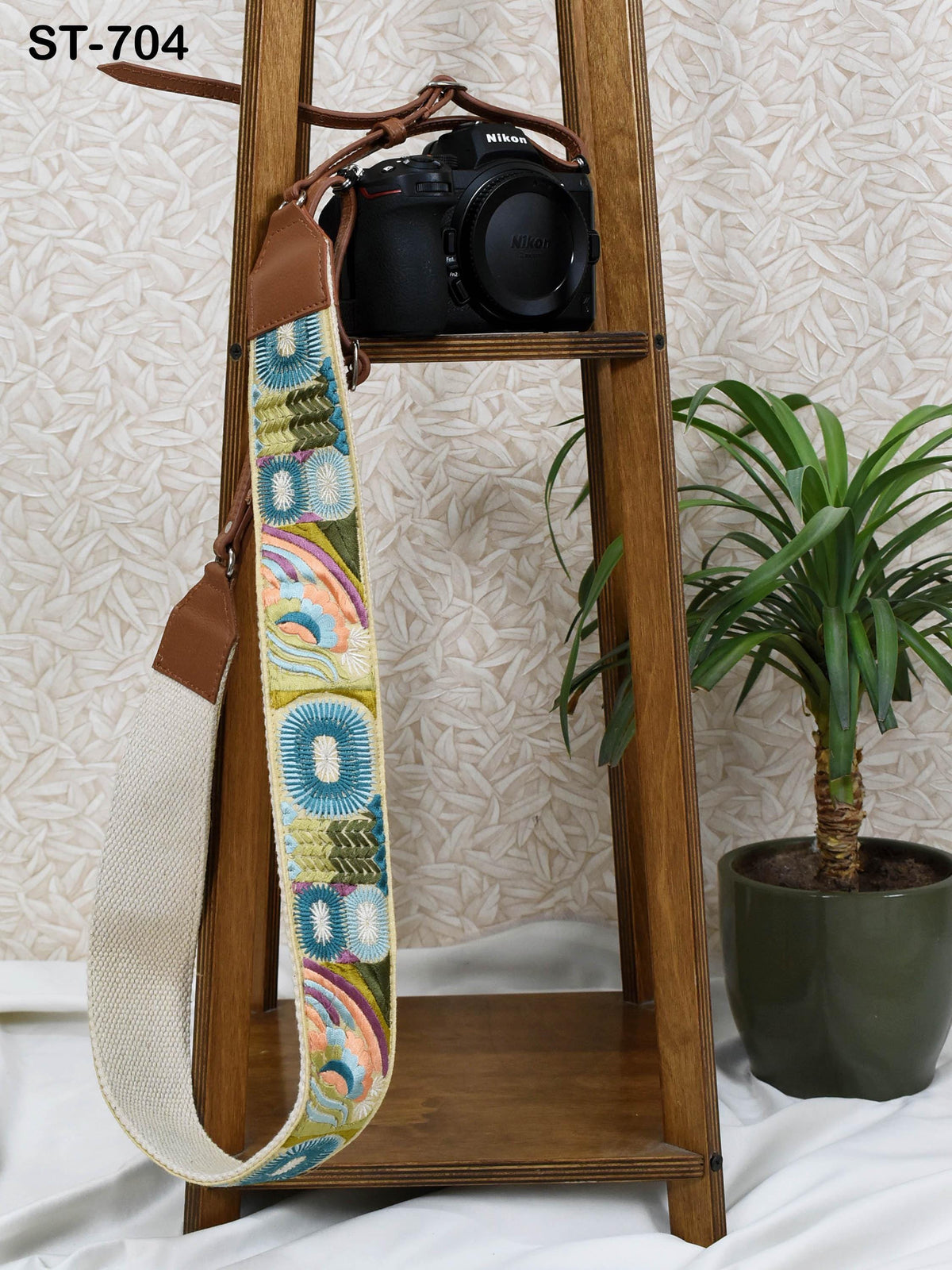 a camera strap with a colorful, patterned design hanging from a wooden stand, next to a potted plant and a camera.