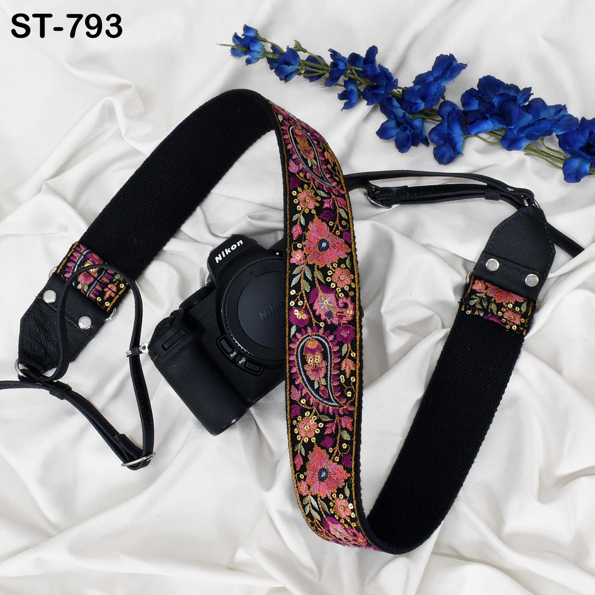 A black camera strap with a colorful floral pattern and a black camera body are placed on a white surface, accompanied by a sprig of blue flowers.