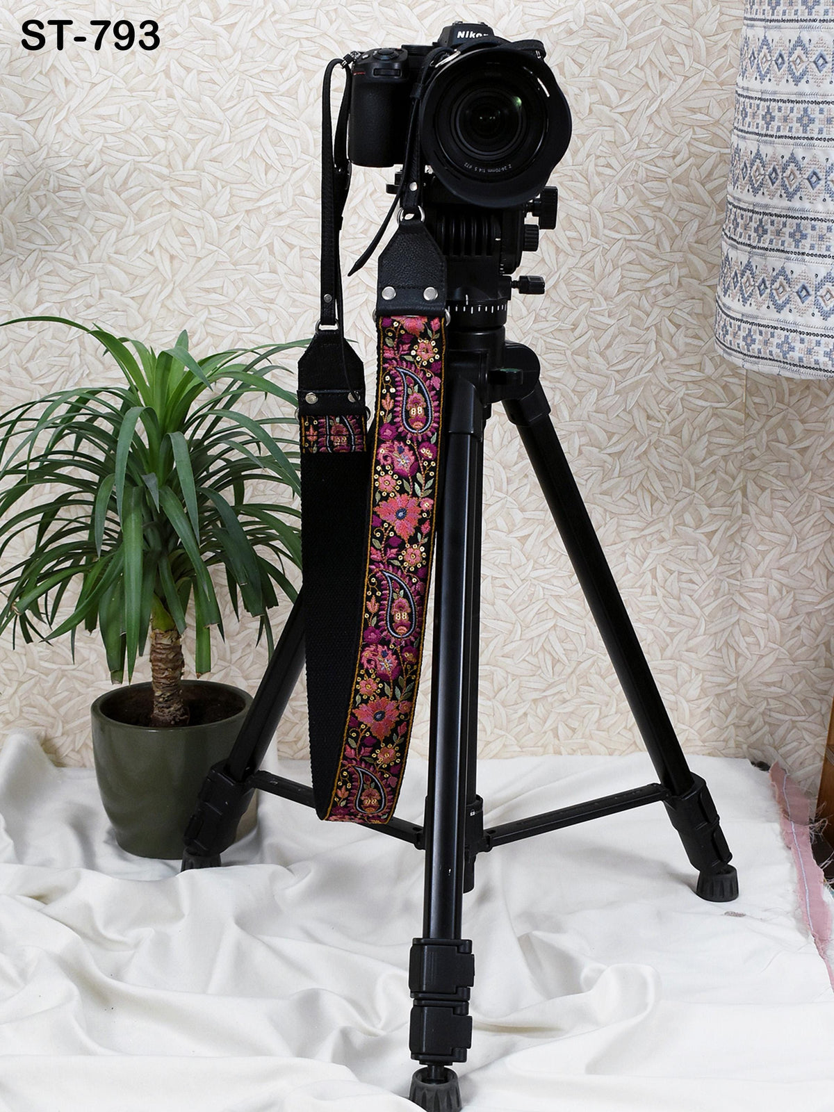 A black tripod with a camera attached to it, placed on a white surface next to a potted plant.