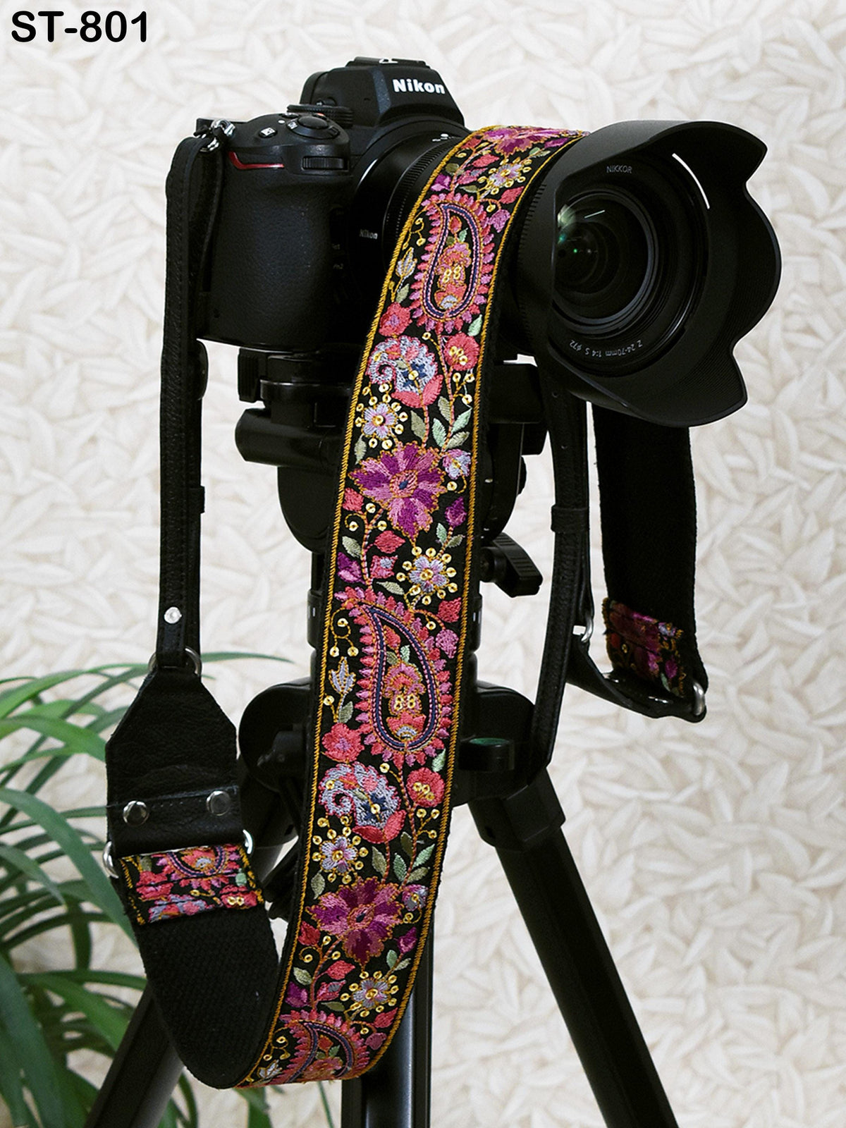 A black camera with a colorful, patterned strap attached to it, placed on a tripod against a white background.