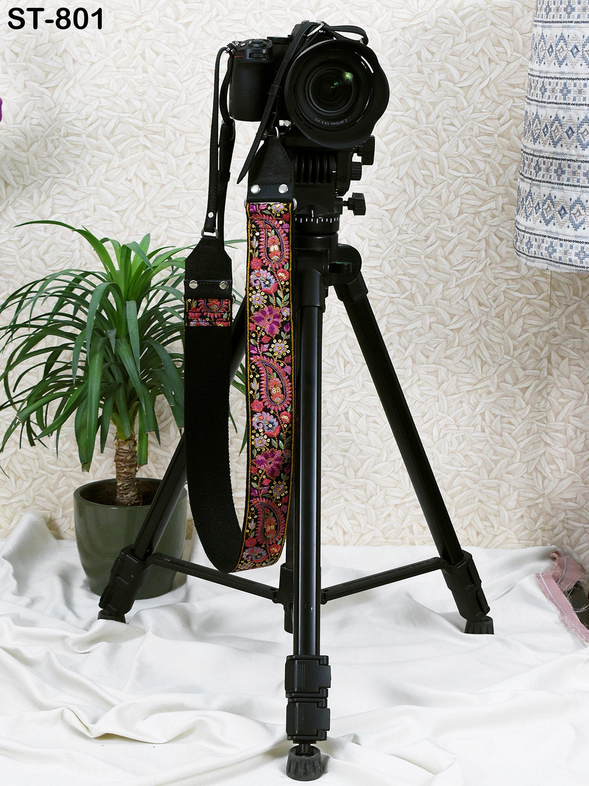 A black tripod with a camera attached to it stands on a white cloth, with a potted plant nearby.