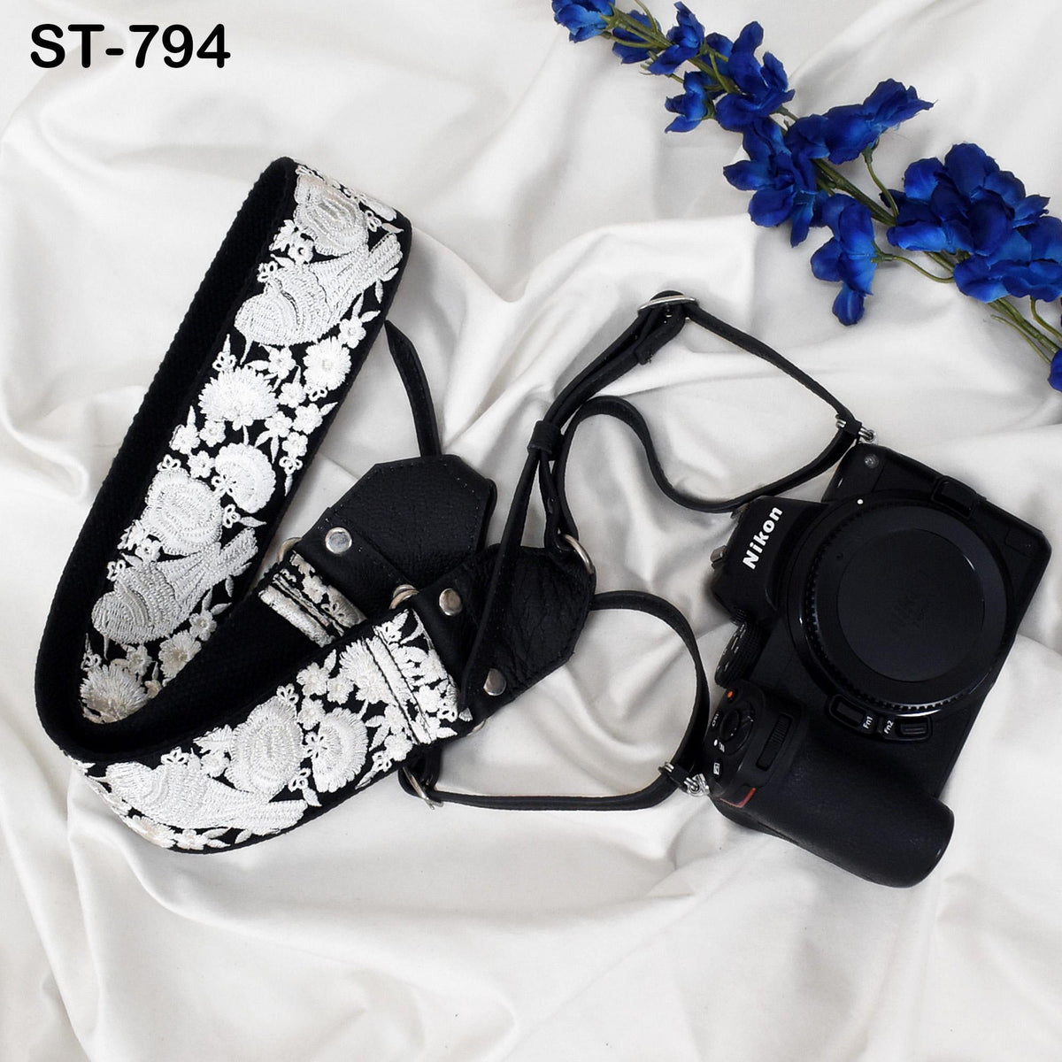 A black and white floral camera strap and a black Nikon camera are placed on a white fabric surface, with a branch of blue flowers in the background.