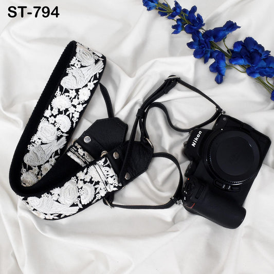 A black and white floral camera strap and a black Nikon camera are placed on a white fabric surface, with a branch of blue flowers in the background.