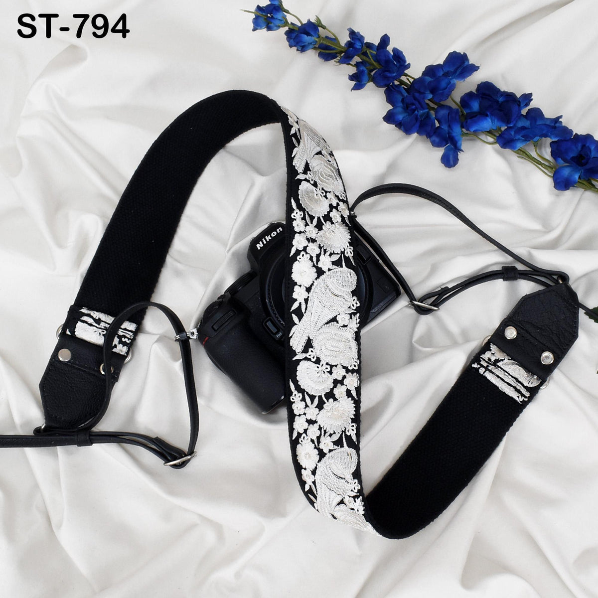 A black camera strap with a white floral pattern is placed on a white fabric surface, accompanied by a bunch of blue flowers.