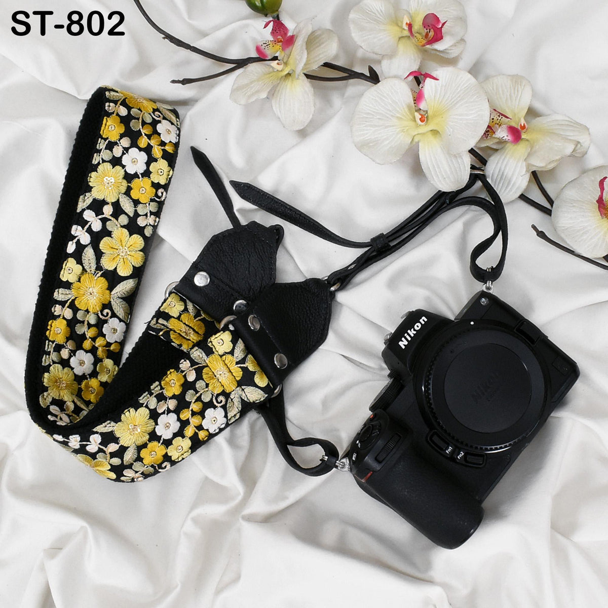 A floral patterned camera strap and a Nikon camera are placed on a white fabric surface, with white orchids in the background.