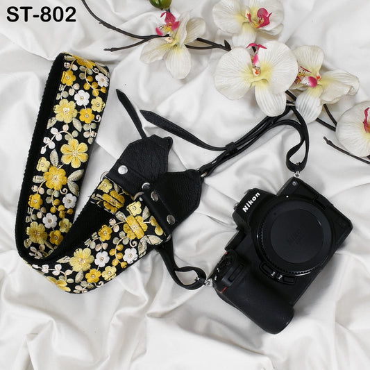 A floral patterned camera strap and a Nikon camera are placed on a white fabric surface, with white orchids in the background.