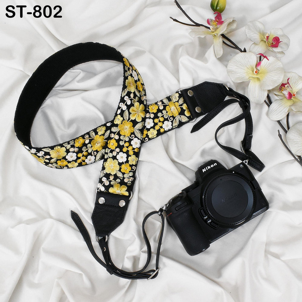 A floral patterned camera strap and a Nikon camera are placed on a white fabric surface, with a few orchids in the background.